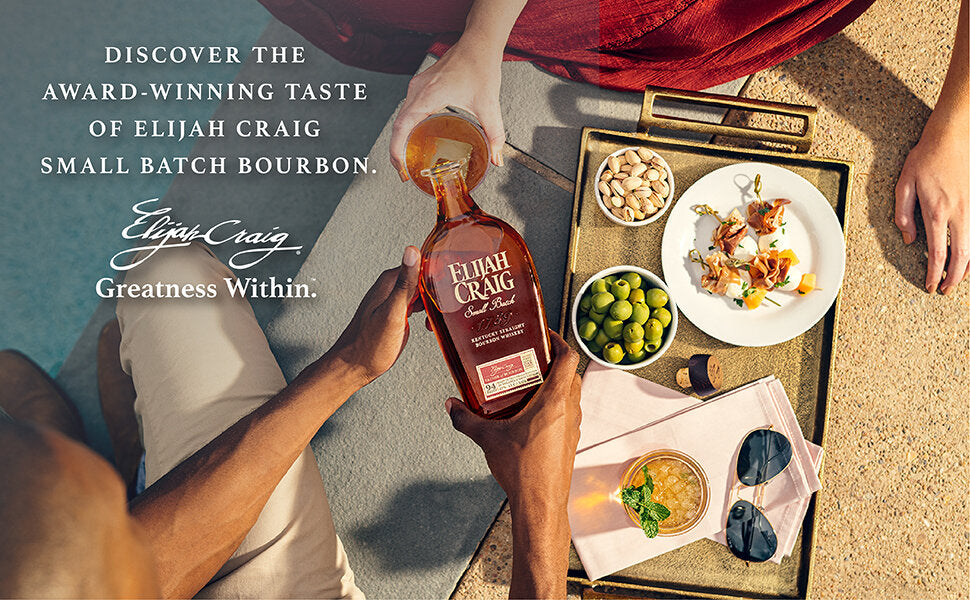 Man pouring bottle of Elijah Craig into woman's rocks glass poolside. Charcuturie style snack tray and cocktail to the right.