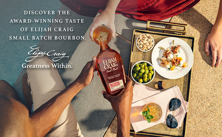 Man pouring bottle of Elijah Craig into woman's rocks glass poolside. Charcuturie style snack tray and cocktail to the right.