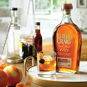 Right, old fashioned cockatil ingredients in background. Left, Eljah Craig small batch bottle with old fashioned on a marble tray.