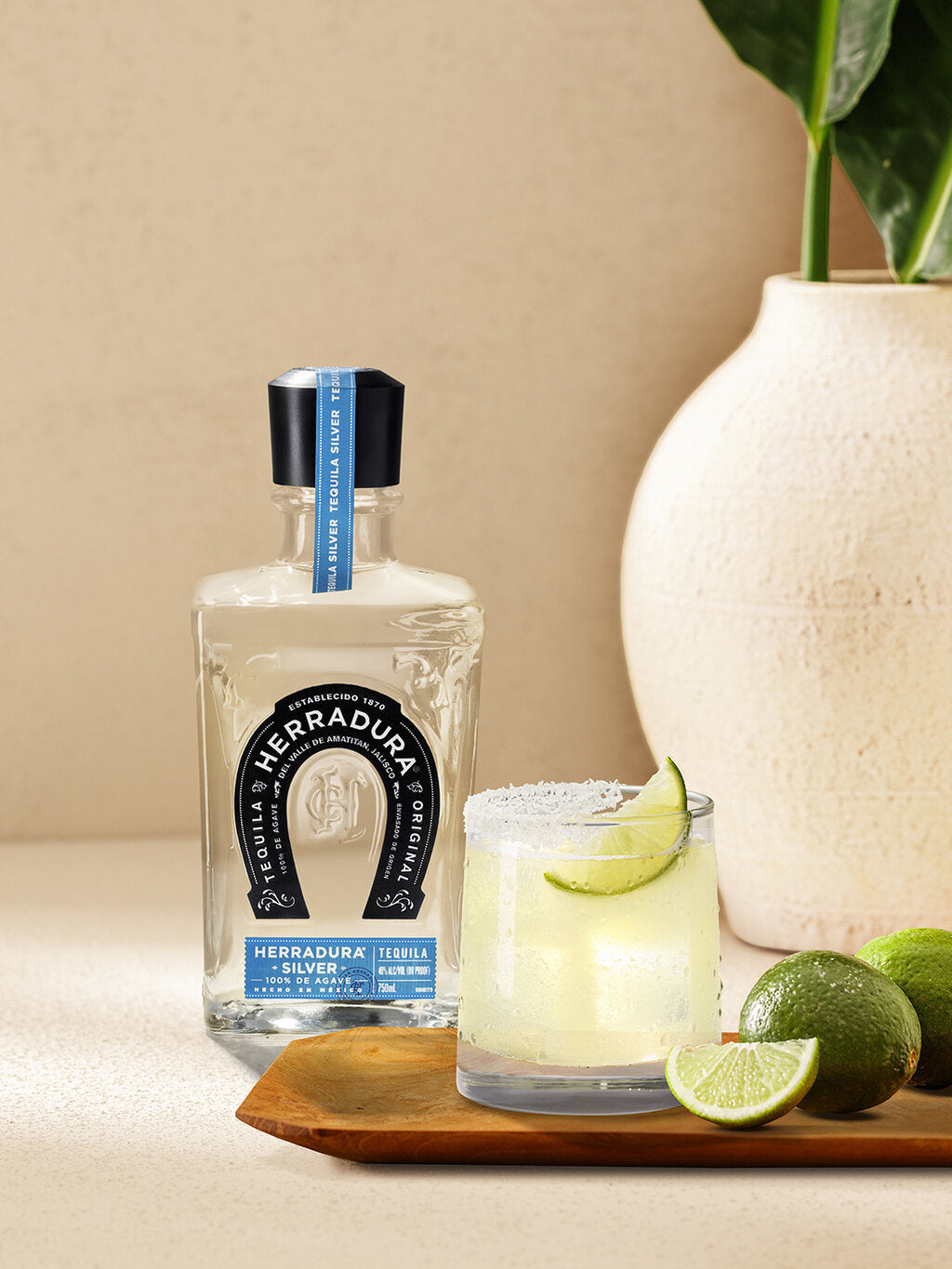 Bottle of Herradura tequila with a HORSESHOE MARGARITA cocktail glass, with lemons on a table.