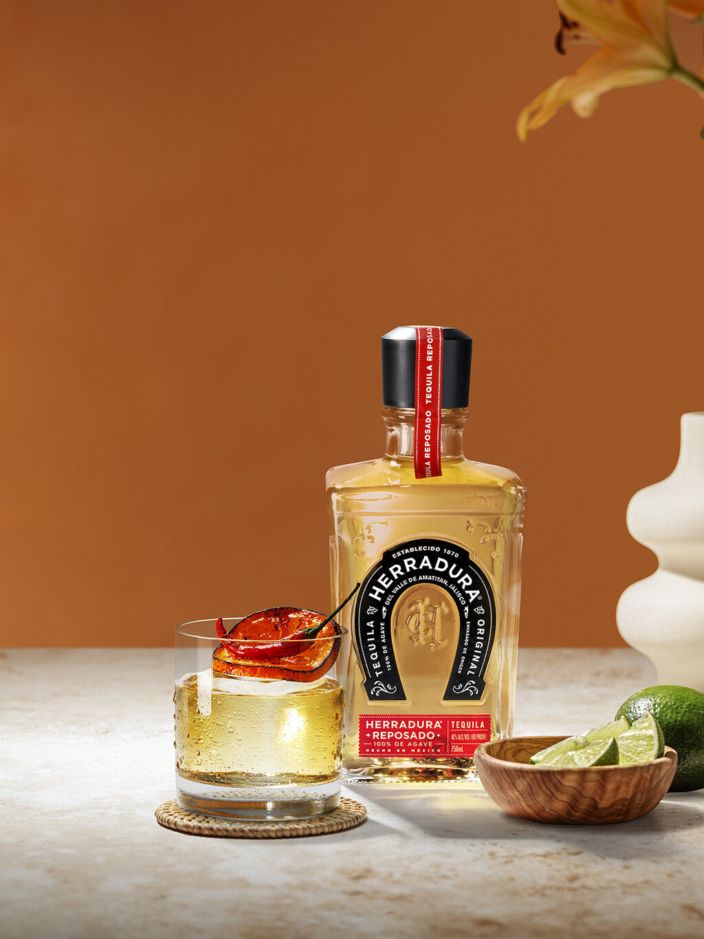 Bottle of HERRADURA REPOSADO Tequila with a glass of SMOKED CHILI OLD FASHIONED cocktail, next to lemons in a small bowl on a table.