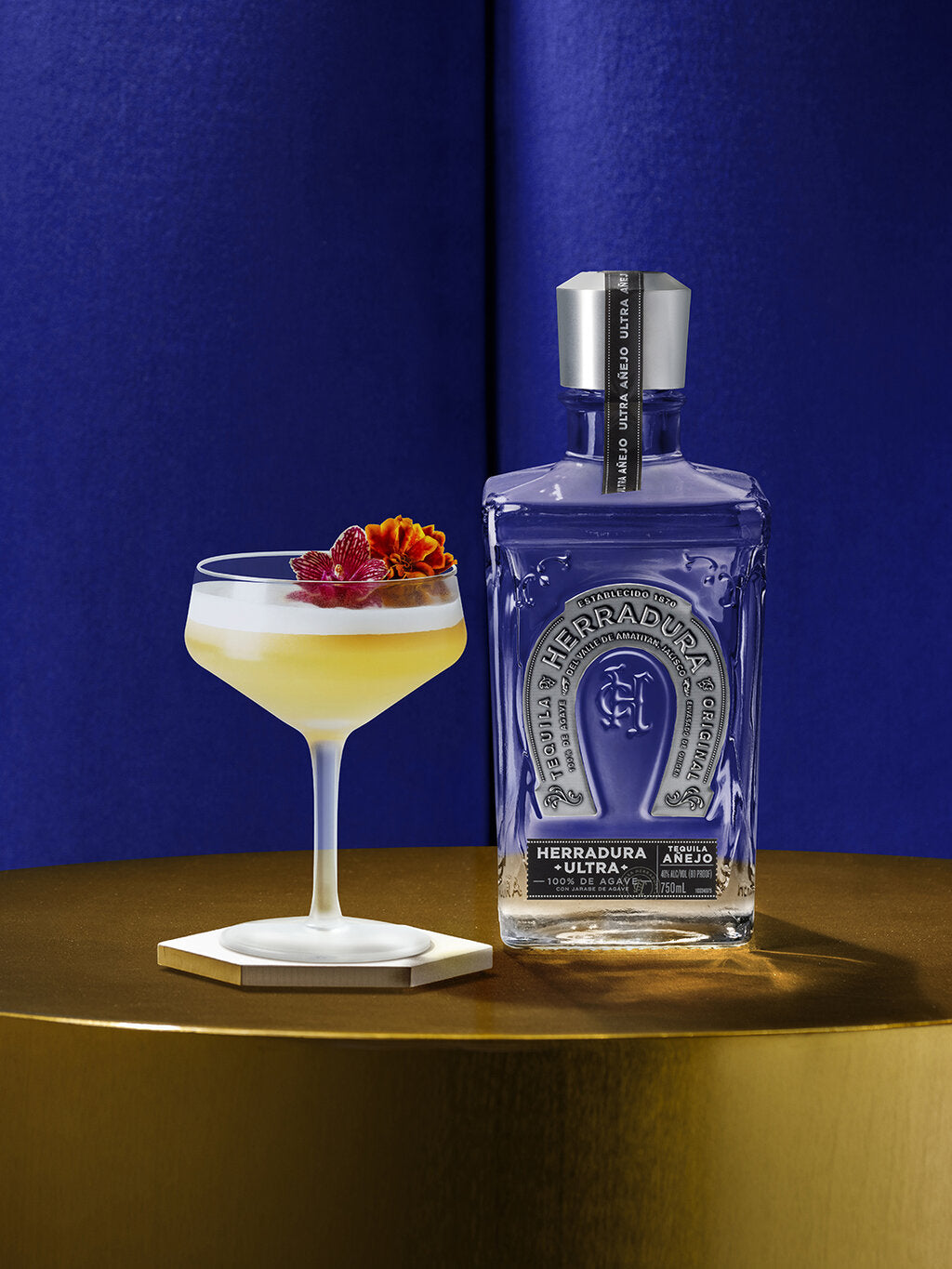 bottle of HERRADURA ULTRA with a glass of APRICOT SOUR cocktail on a gold table.